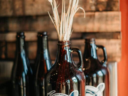 A Badger State Brewing glass growler filled with wheat stalks sits in front of two dark glass bottles on a wooden surface.
