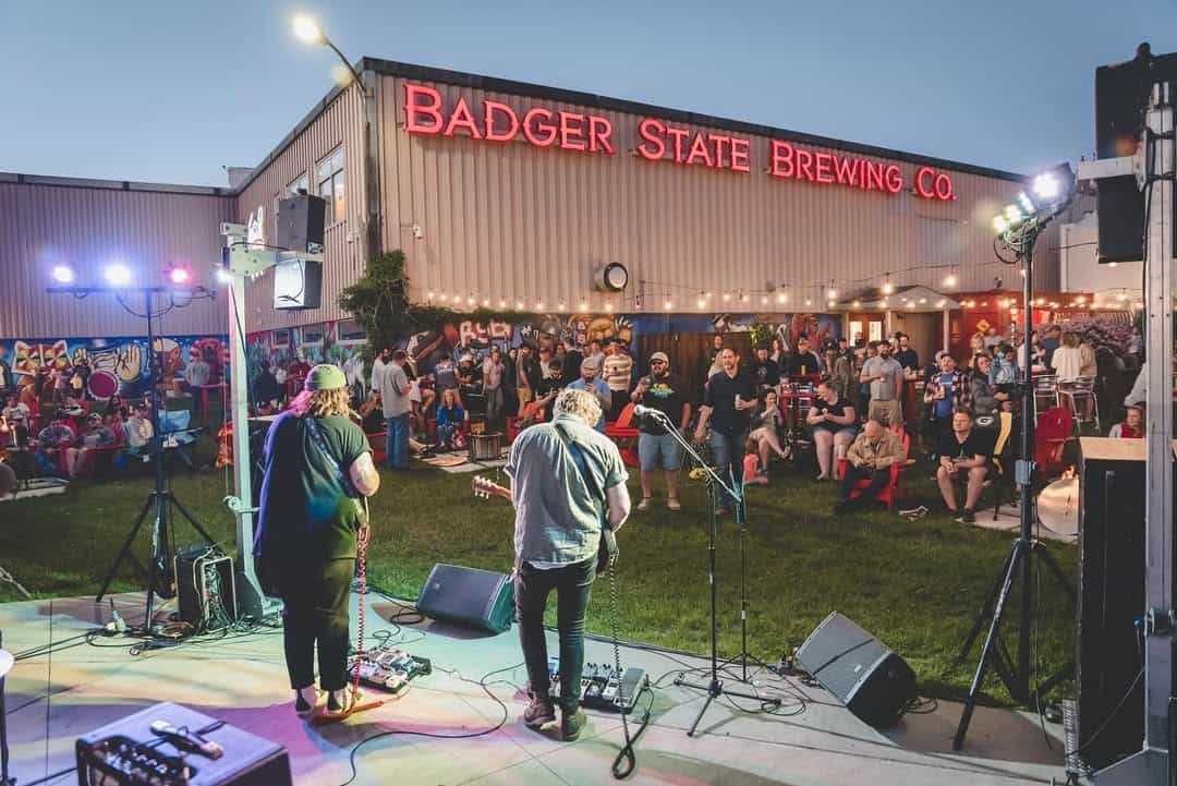 A band plays on an outdoor stage at Badger State Brewing Co. in the evening as a crowd gathers on the lawn beneath the lit brewery sign.