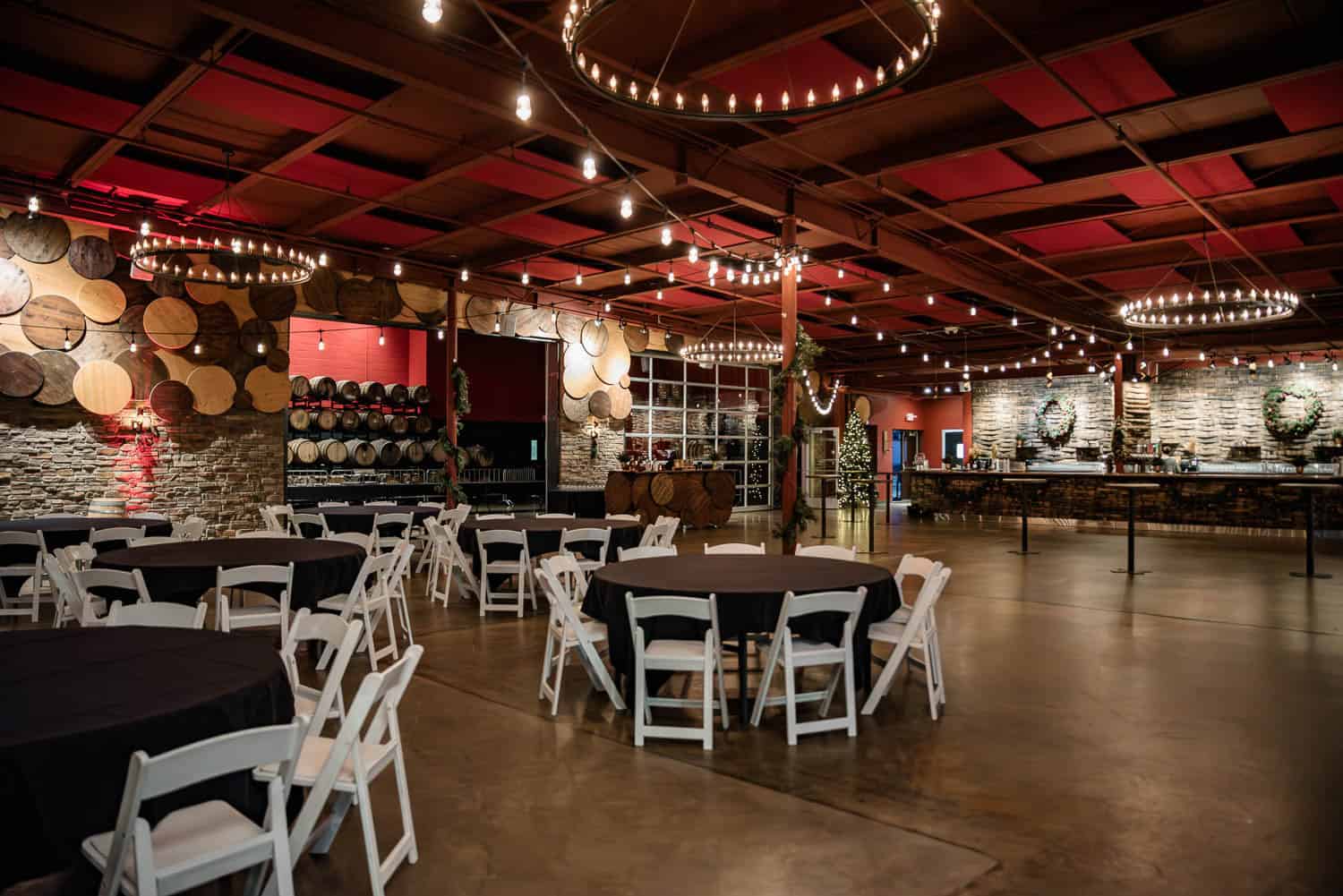 BSB-Events-10 Spacious event hall with round tables and white chairs, wine barrels on the walls, chandeliers, and a bar area in the background.