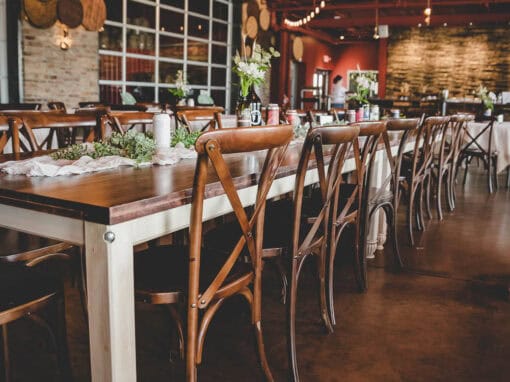 A rustic dining area with wooden tables and chairs, decorated with small floral arrangements, under circular chandeliers and warm lighting.