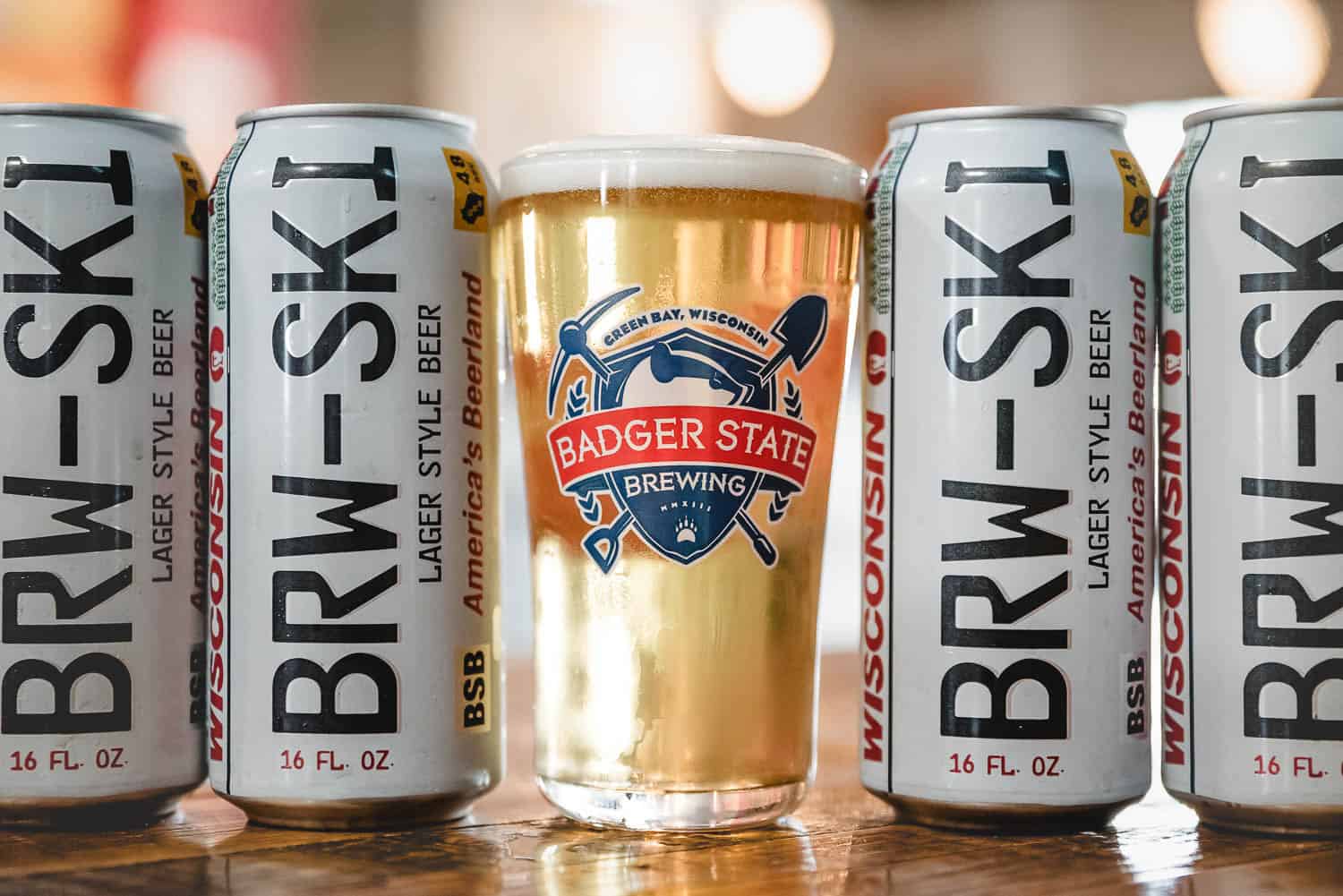 Badger-State-Brewing-Events-242 Four cans of BRW-SKI Lager Style Beer surround a pint glass filled with beer, featuring the Badger State Brewing logo.
